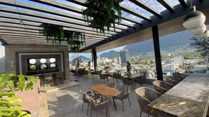 Area salotto esterna con vista sulla città, caratterizzata da un tavolo e sedie in legno, un barbecue e piante pensili.