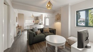 Una sala de estar moderna con un sofá, una mesa de centro y un televisor. Se observa una cocina al fondo, con un ventanal.
