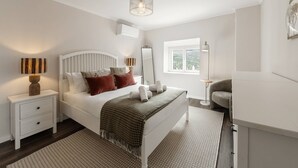 Un dormitorio con cama blanca, mesita de noche, lámpara, espejo y ventana.