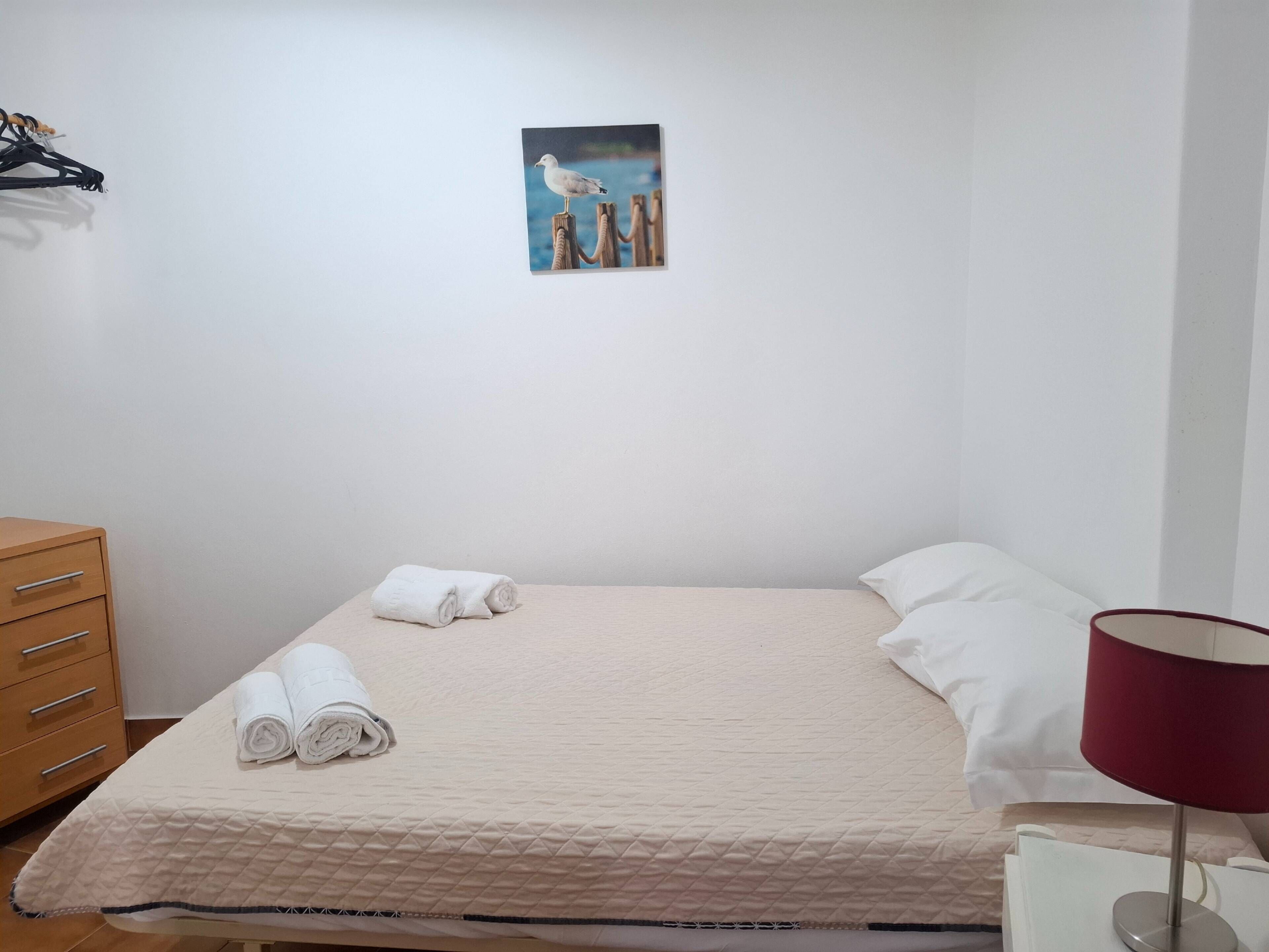 Un lit bien fait, recouvert d’une courtepointe matelassée, une pile de serviettes enroulées, une table de chevet en bois avec une lampe rouge, et un tableau représentant des mouettes accroché au mur.