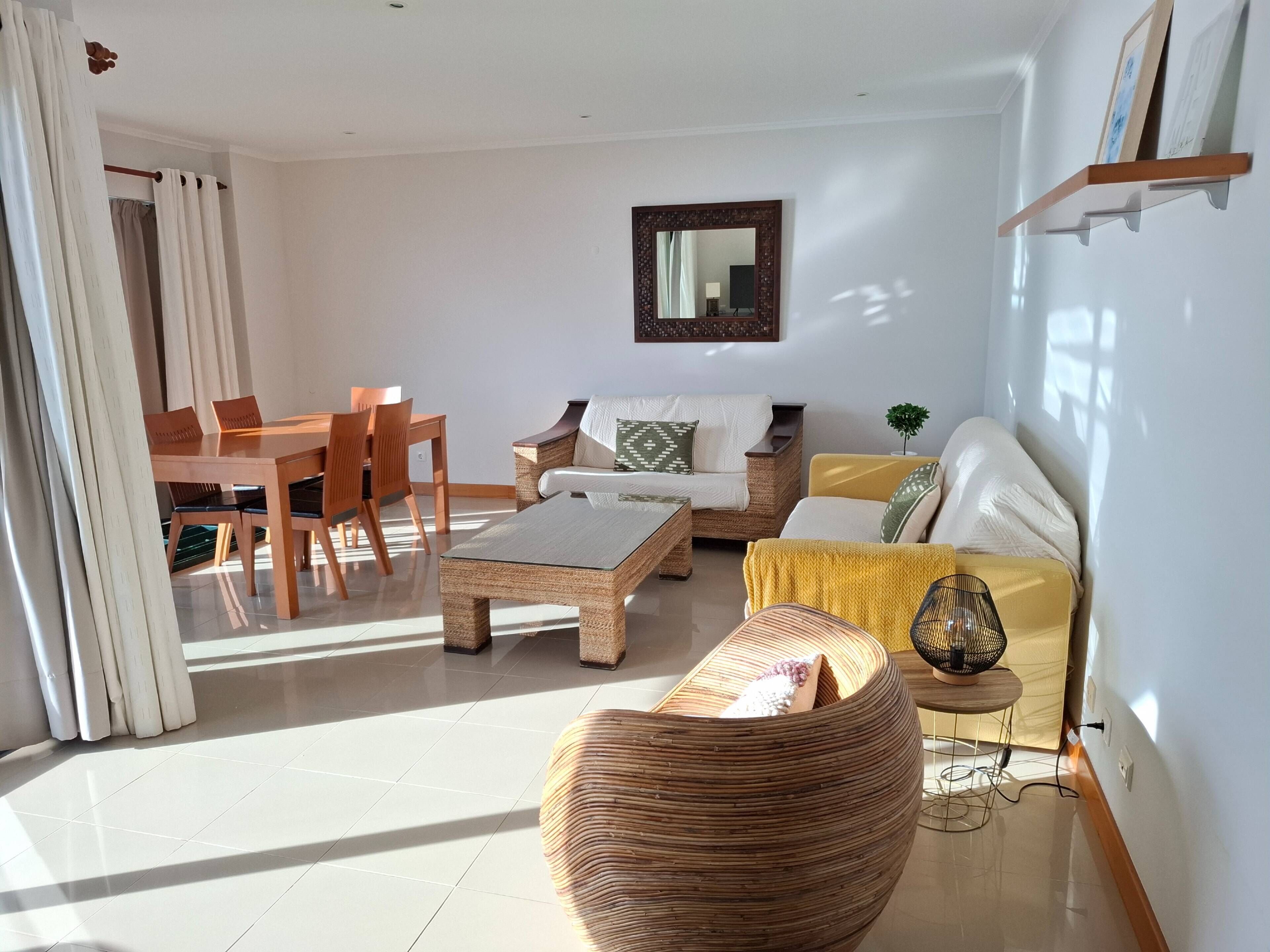 Sala de estar con sofá, mesa de centro y sillas. Se observa una mesa de comedor con sillas. Una planta pequeña está sobre una mesita auxiliar.