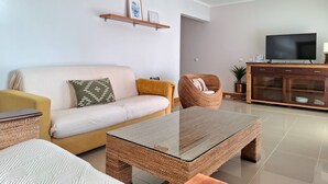 A modern living room with a sofa, a glass coffee table, a wicker chair, a TV on a wooden cabinet, and a small shelf with pictures. - PRAIA DA ROCHA- CASA DA PRAIA 904 (Portimão)