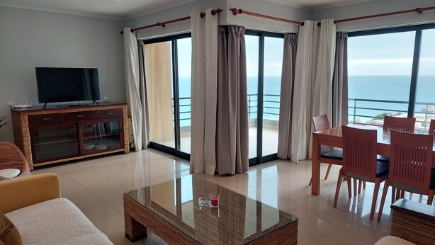 A spacious living room with a large glass coffee table, comfortable seating, a TV on a wooden cabinet, and a balcony with a view of the sea. - PRAIA DA ROCHA- CASA DA PRAIA 904 (Portimão)