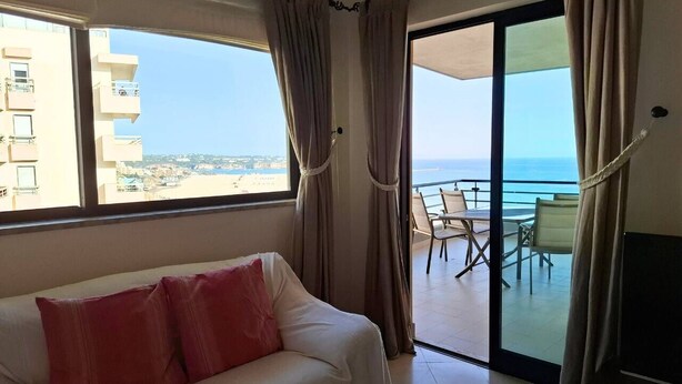 A hotel room with a white sofa, a balcony with a table and chairs, and a view of the sea and buildings. - PRAIA DA ROCHA- CASA DA PRAIA 702 (Portimão)