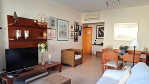 A living room with a flat-screen TV, wicker furniture, a white sofa, and a dining area with a table and chairs. - PRAIA DA ROCHA- CASA DA PRAIA 702 (Portimão)