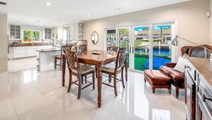 Eine geräumige Küche mit weißen Schränken, ein Essbereich mit einem Holztisch und -stühlen sowie Blick auf einen Pool durch Glastüren.