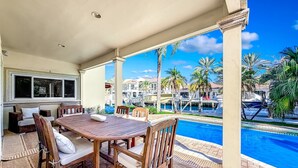 Ein überdachter Außenbereich mit Pool, Holzmöbeln und Blick auf Boote und Palmen.