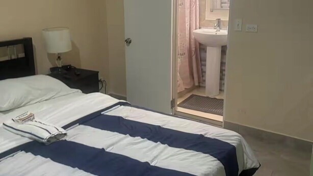 Hotellihuone, jossa on sänky, yöpöytä lampun kanssa ja kylpyhuone, joka näkyy avoimesta ovesta.