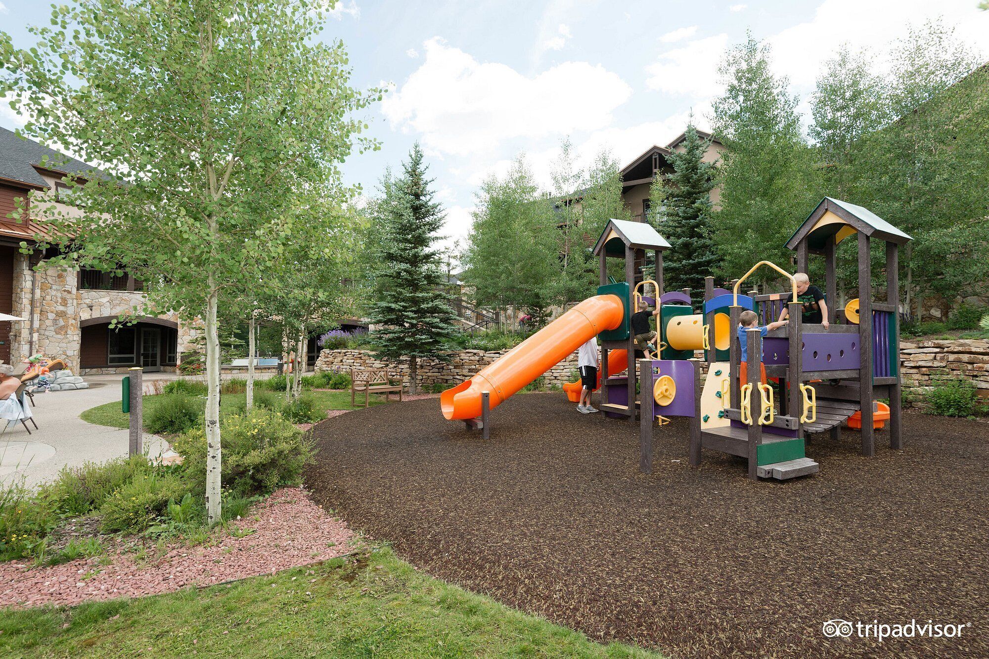 Un parque infantil con tobogán y estructuras para escalar, rodeado de árboles y un muro de piedra.
