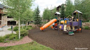 A playground with a slide and climbing structures, surrounded by trees and a stone wall. - Beautiful Family Friendly Luxury Resort - 2.20.26 to 2.27.26 - Tons of Amenities (Breckenridge)