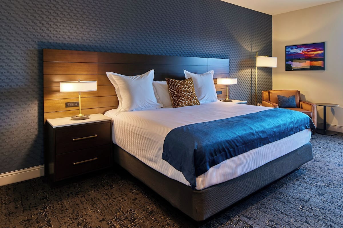 A modern hotel room with a large bed, two bedside tables with lamps, a wooden headboard, and a view of a sunset.