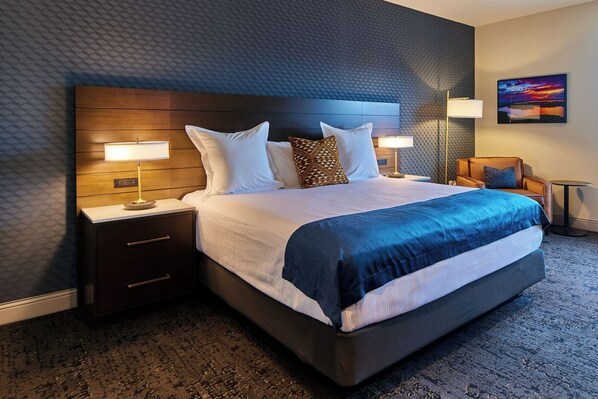 Una habitación de hotel moderna con una cama grande, dos mesitas de noche con lámparas, un cabecero de madera y vistas a un atardecer.