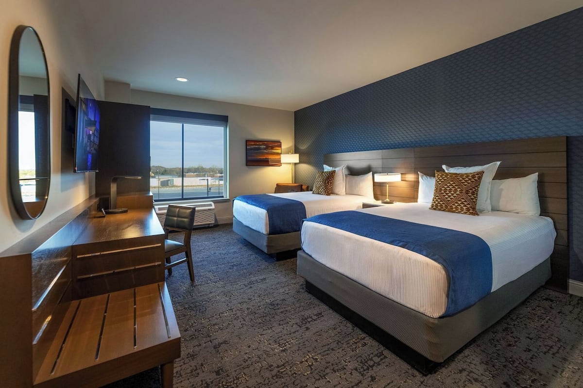 A hotel room with two beds, a desk, a chair, a TV, and a large window with a view.