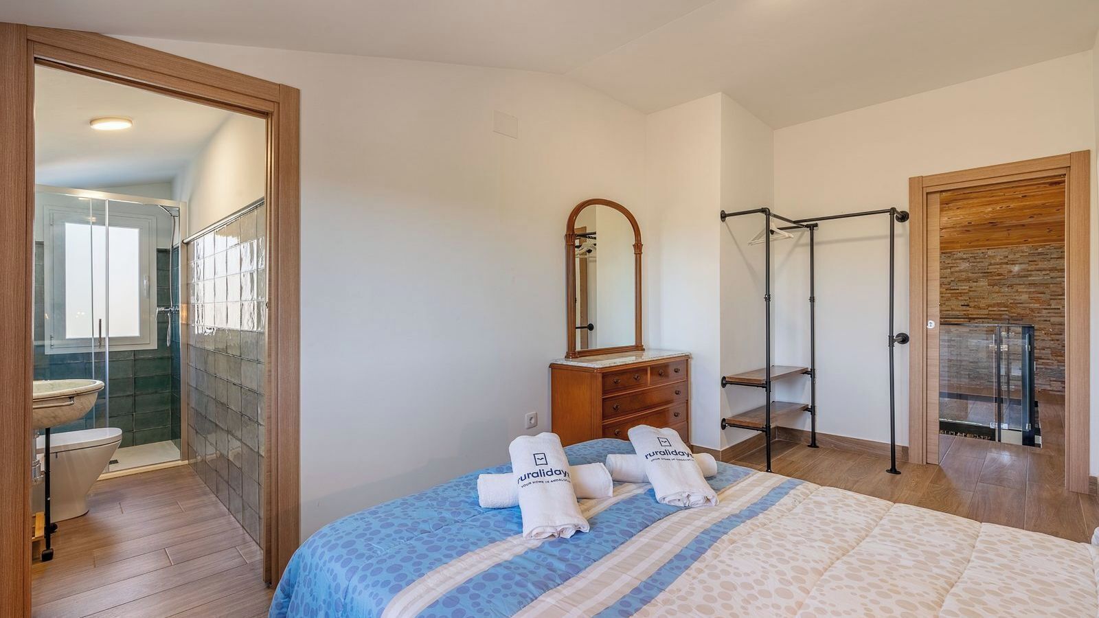 A hotel room with a bed, a wooden dresser, a mirror, a towel rack, and a bathroom with a shower and toilet.