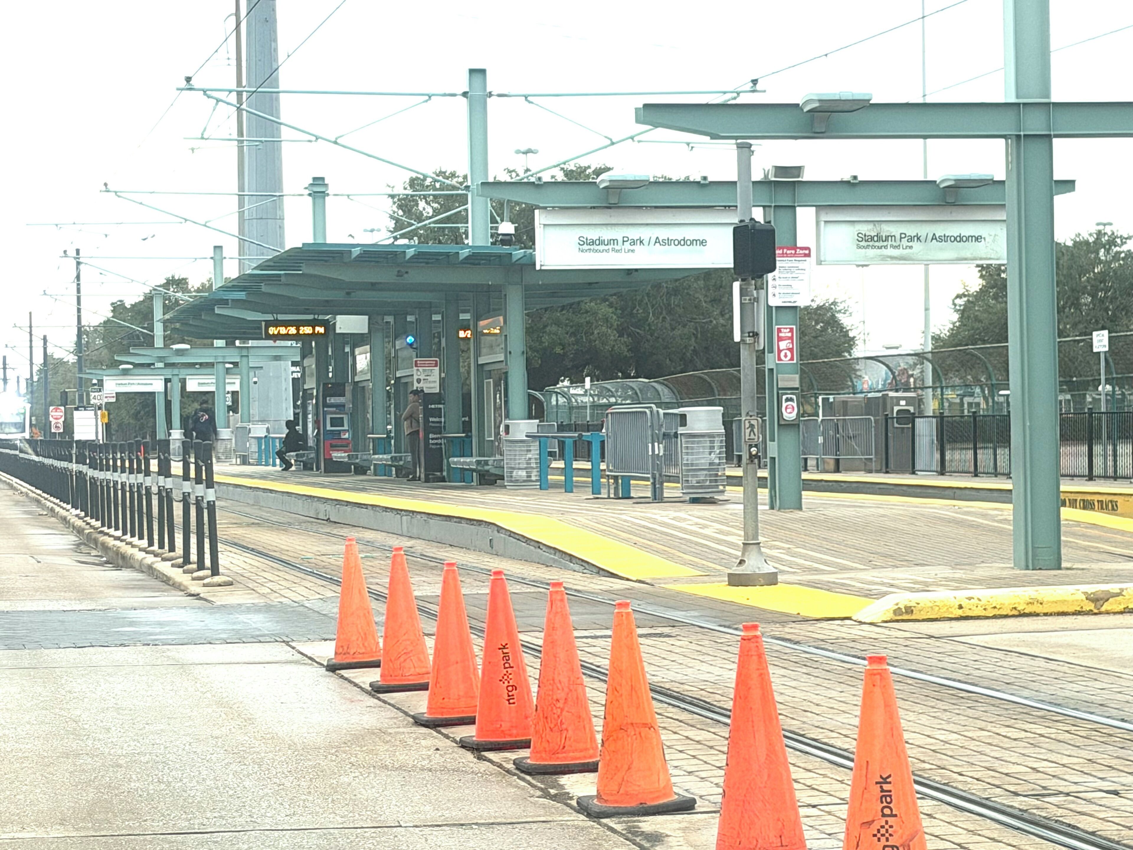 Una banchina di stazione ferroviaria con coni stradali arancioni, un'area di attesa coperta e indicazioni per Stadium Park e Astrodome.