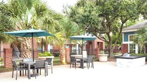 Patio esterno con tavoli e sedie, immerso tra alberi e affiancato da un edificio in mattoni.