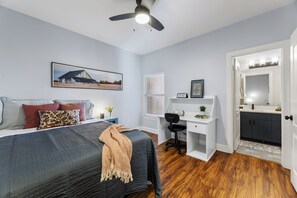 Un dormitorio con cama, escritorio, silla, ventilador de techo, ventana con persianas y baño con lavabo y espejo.
