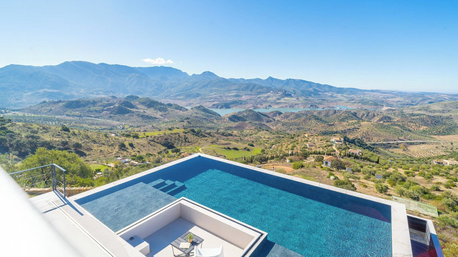 Una piscina d'hidromassatge rectangular de forma infinita, amb aigua cristal·lina blava, envoltada per un solàrium blanc i una petita zona de seients amb taula i cadires.