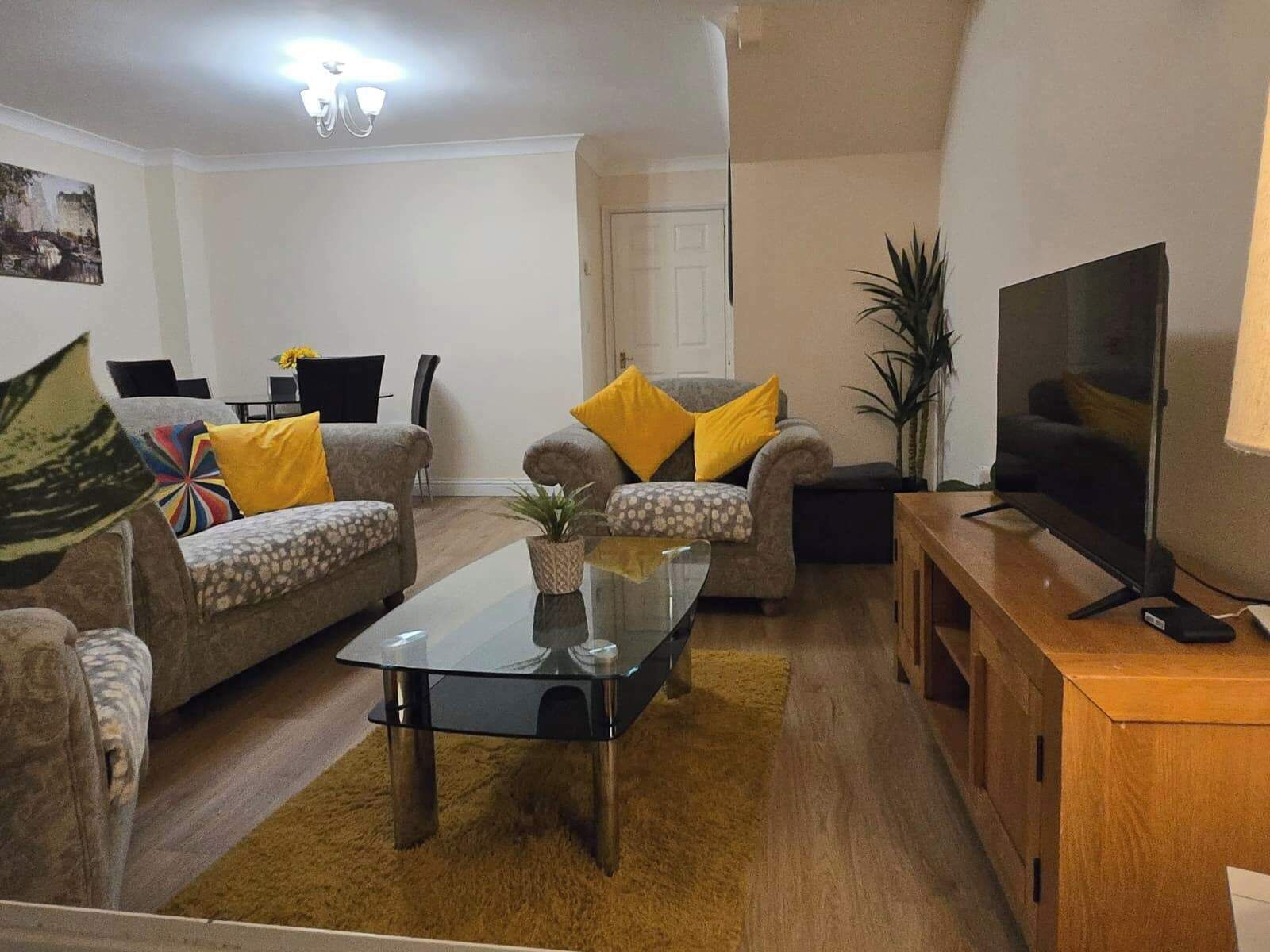 Ein Wohnzimmer mit zwei Sofas, einem Glastisch, einem Fernseher auf einem Holzständer und einer Tür im Hintergrund.