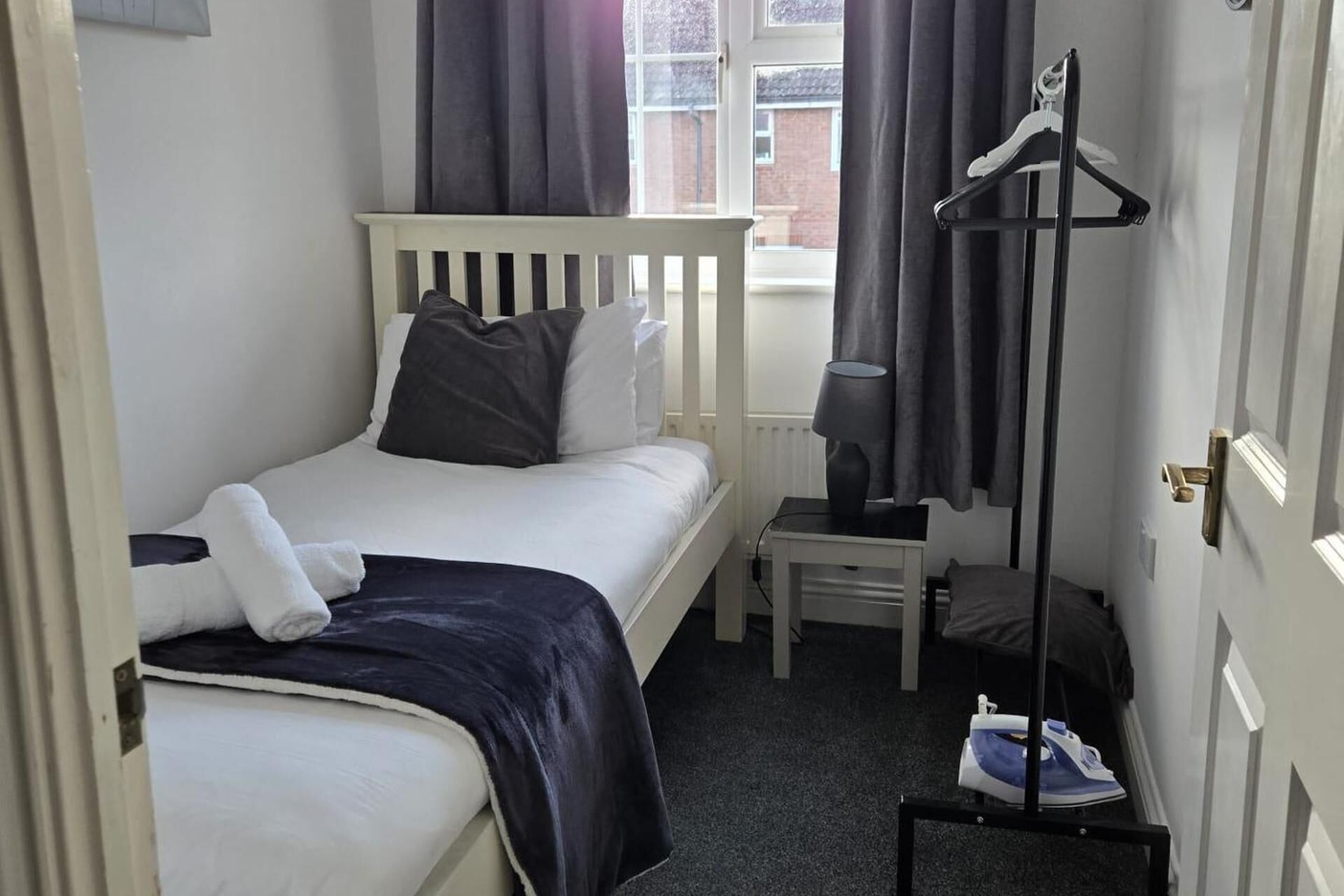 A small, neatly arranged bedroom with a single bed, a bedside table with a lamp, a hanging clothes rack, and a small ironing board.
