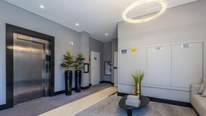 Un hall d’entrée moderne équipé d’un ascenseur, de plantes en pot, d’un comptoir de réception et d’un panneau indiquant un espace d’attente général.