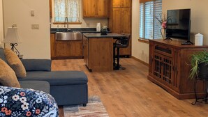 Una sala de estar moderna con un sofá, una mesa baja y un televisor. Se observa un área de cocina con gabinetes de madera y un lavamanos.