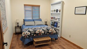 Un dormitorio con cama, mesita de noche, estantería y una ventana con persianas. Hay una cita enmarcada en la pared.