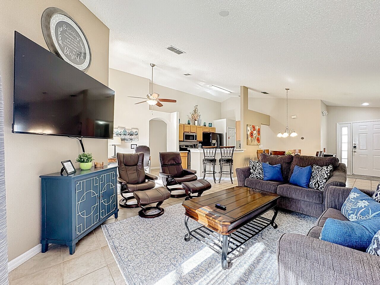 A spacious living room with a flat-screen TV, comfortable seating, a coffee table, and a kitchen area in the background.