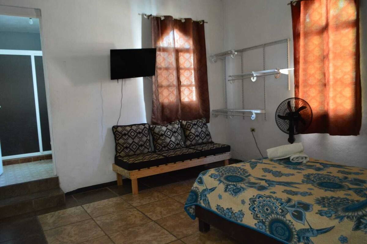 A bedroom with a bed, a sofa, a TV mounted on the wall, a fan, and a window with curtains.