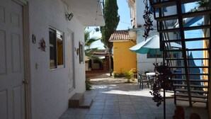 Une cour étroite avec un sol carrelé, un bâtiment blanc doté d’une porte et de fenêtres, un bâtiment jaune en arrière-plan, et des sièges extérieurs avec des parasols.