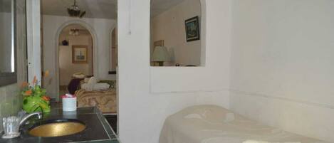 A hotel room with a bathroom featuring a sink, mirror, and green-tiled vanity. There is a bed with a white comforter and a small table with a lamp.