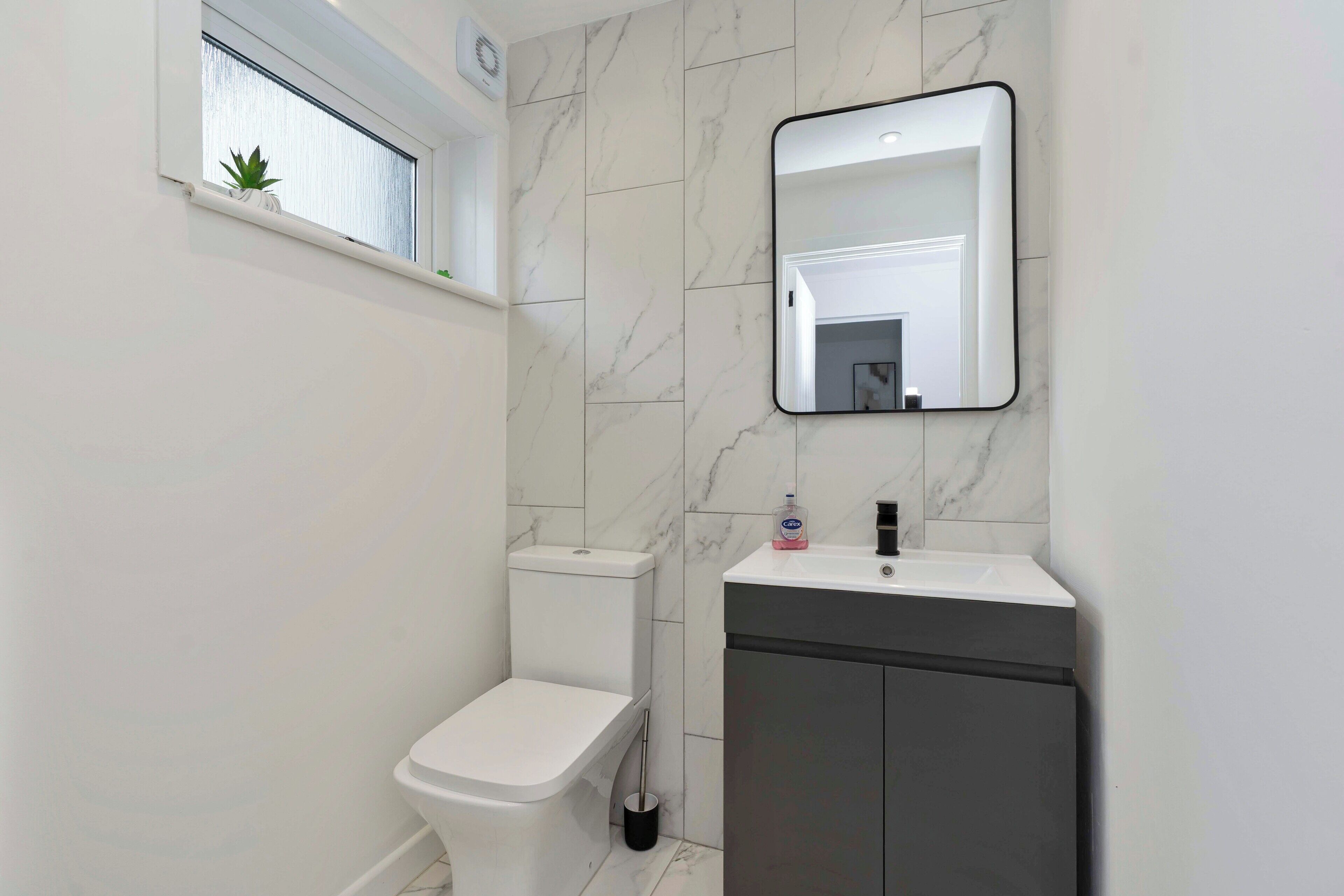 Un baño moderno con inodoro, lavamanos, espejo y ventana con planta.