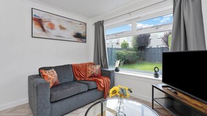 A modern living room with a grey sofa, a coffee table with a vase of flowers, a flat-screen TV on a wooden stand, and a large window with curtains. - Stylish 4bed Home Free Parking 20 Min to Edinburgh (Livingston)