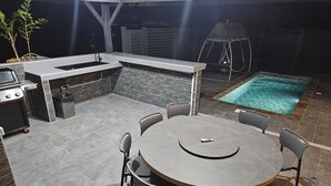 Een terras buiten met een ronde tafel en stoelen, een barbecue, een klein zwembad en een schommelset.