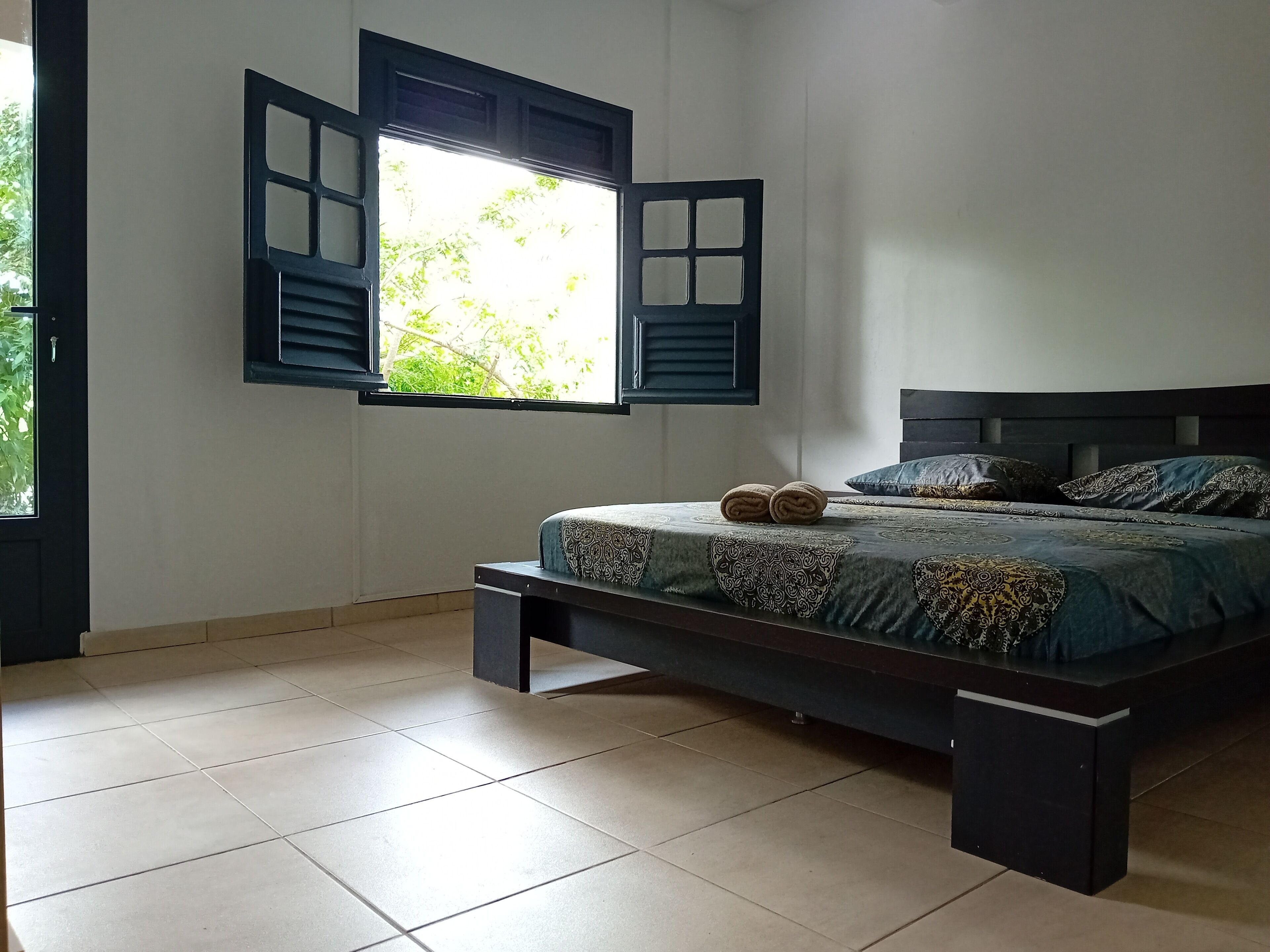 Un dormitorio con cama, ventana con contraventanas abiertas y vistas a zonas verdes.