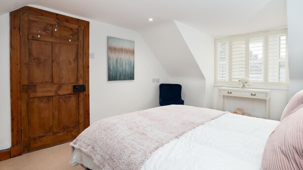 Een slaapkamer met een grote houten deur, een bed met een roze dekbed, een blauwe stoel, een witte ladekast en een raam met zonwering.