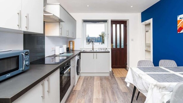 A modern kitchen with a microwave, oven, and sink. There is a dining area with a table and chairs. The room has a window and a view of the outside. - Airport Apartment - Aberdeen - Free Parking (Aberdeen)