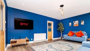 A living room with a blue wall, a flat-screen TV, a blue sofa with orange cushions, a wooden coffee table, and a patterned rug. - Airport Apartment - Aberdeen - Free Parking (Aberdeen)