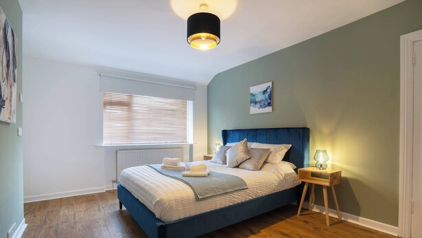 Ein Schlafzimmer mit Bett, Nachttischen, einem Fenster mit Jalousien, einem Heizkörper und einer Deckenleuchte.