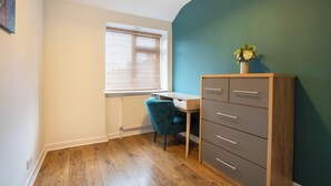 Ein Zimmer mit einem Holztisch, einem blauen Stuhl, einer grauen Kommode und einem Fenster mit Jalousien.