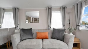 Un salón con tonos neutros, un sofá gris, dos cojines, un espejo y vistas a una zona residencial por la ventana.