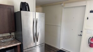 Eine kompakte Küche mit einem Edelstahl-Kühlschrank, einem Holflanschrank, einer schwarzen Mülltonne und einem an der Wand montierten Feuerlöscher.