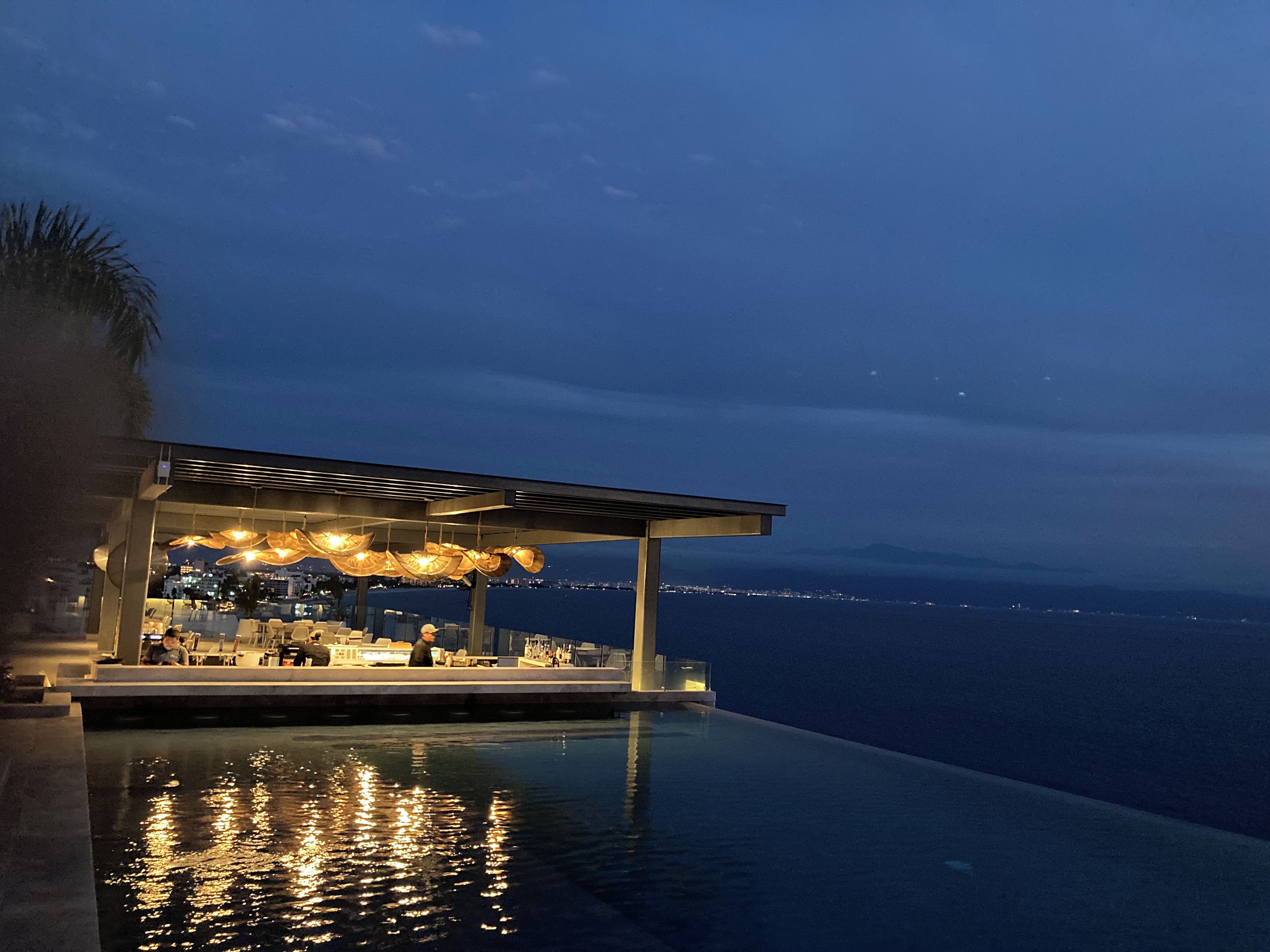 Une piscine à débordement offrant une vue sur la mer, un espace extérieur couvert avec des éclairages suspendus et un design architectural moderne.