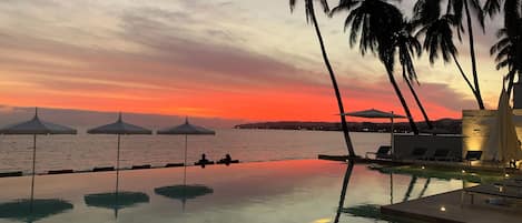 Zona de piscina con sillas de descanso y sombrillas, con vista a un cuerpo de agua durante el atardecer.