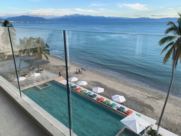 Piscina con sillas de descanso y sombrillas, con vistas a una playa y un paisaje montañoso.
