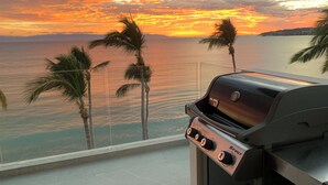 Un balcón con una parrilla que da a una vista del atardecer sobre el océano y palmeras.