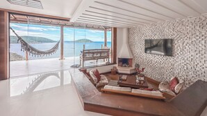 A spacious living room with a large glass door leading to a balcony, a fireplace, and a mounted TV. - Luxury Oceanfront Villa with Infinity Pool, Sauna & Total Privacy (Arraial do Cabo)