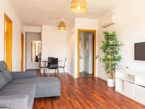 Ein modernes Wohnzimmer mit einem grauen Ecksofa, einem Essbereich mit Tisch und Stühlen, einer großen Kühlpflanze und einem Wand-Fernseher.