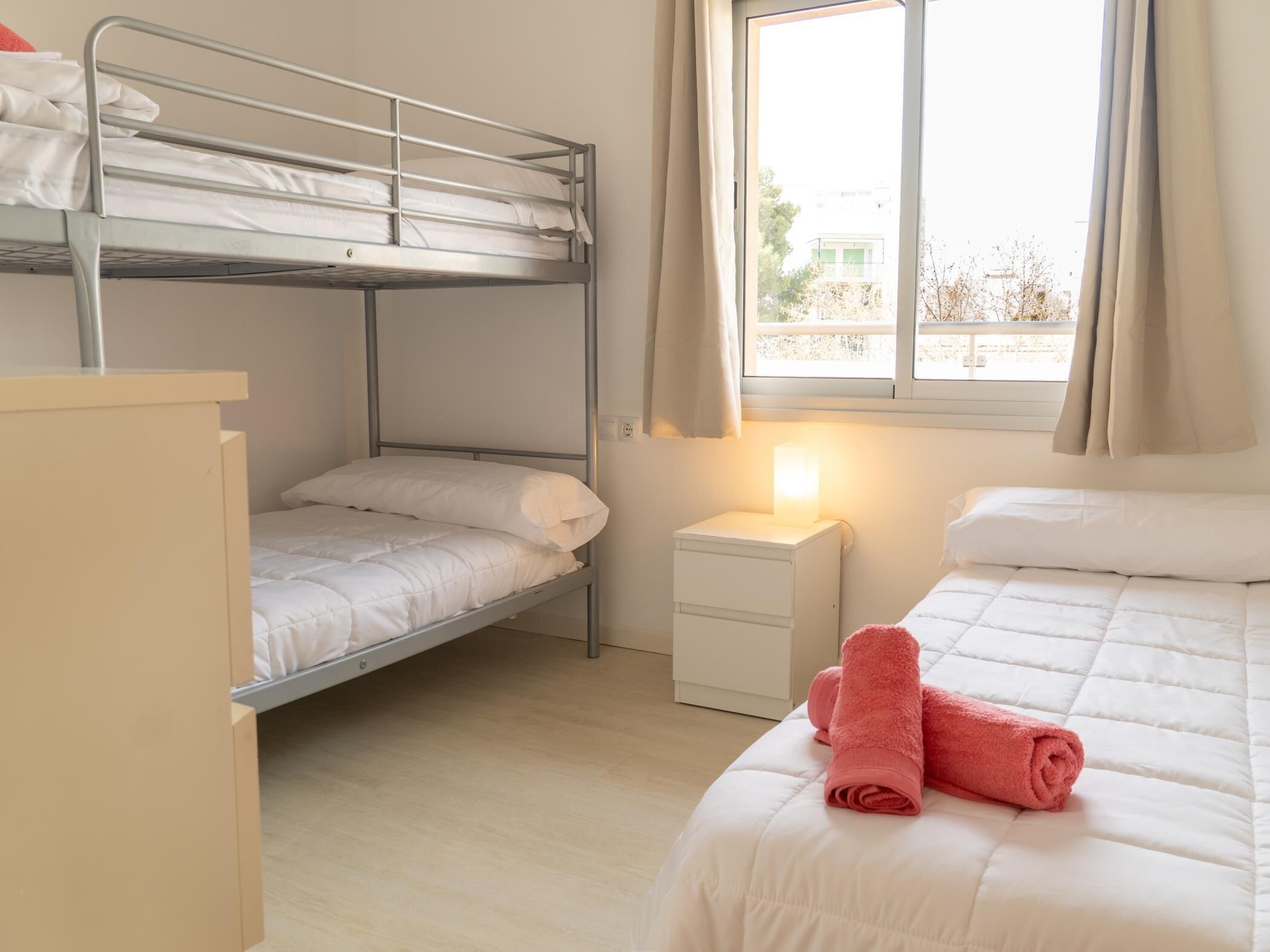 Een slaapzaal met stapelbedden, een nachtkastje met lamp en een raam met gordijnen.
