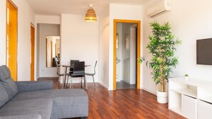 Uma sala de estar moderna com um sofá seccional cinza, uma área de jantar com mesa e cadeiras, uma planta grande em vaso e uma TV na parede.